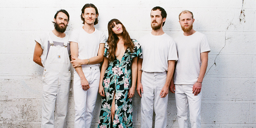 A photo of Amo Amo on a white background, with the lead singer in a bright dress surrounded by the other band members dressed in all white.